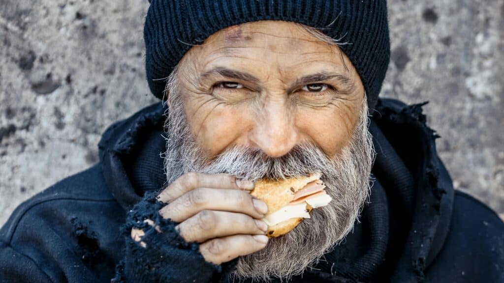 homeless man squatting eating sandwich