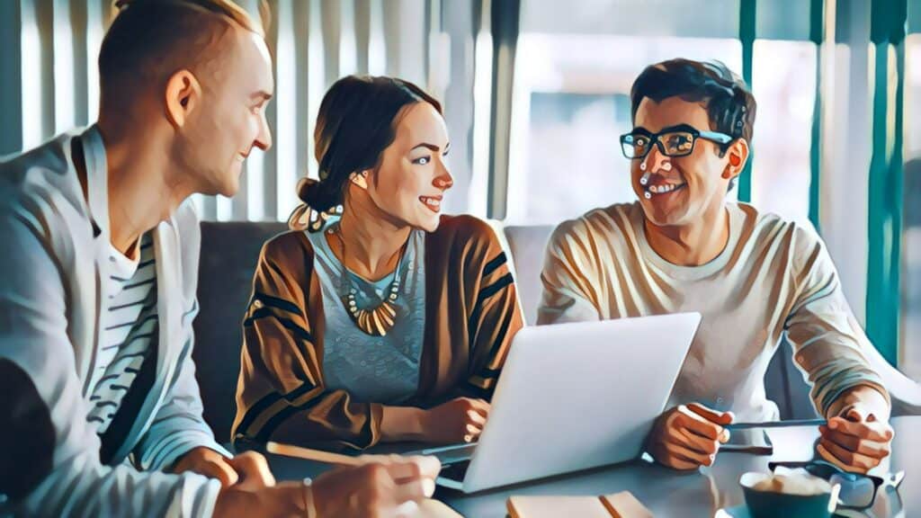 Three people talking and smiling at a small table with laptop computer, man wearing glasses, dark-rimmed, smiling women wearing brown shawl in middle of two men, large picture windows open in background of office setting