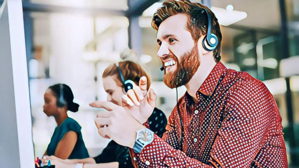 Top 10 Best Ways Virtual Assistants Help Real Estate Investors Increase Cash Flow, man wearing telephone headset, sitting at desk looking and smiling into a computer monitor, wearing a watch, burgundy polka dot long-sleeved dress shirt, in office setting, other workers in the background